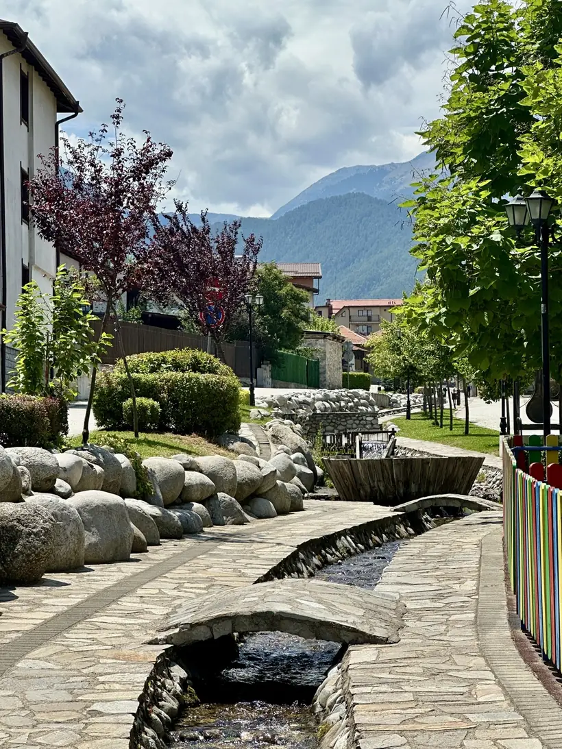 A stone walkway following a mountain stream through Bansko, with a brightly painted children's fence and the Pirin mountains beyond